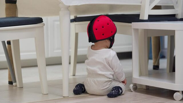Baby wearing soft safety helmet learning to walk, craw sit and stand on the floor in the kitchen, head protector helps reduce impact of falls on hard surfaces. High quality 4k footage