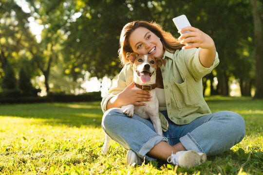 Young caucasian woman student female pet owner taking selfie photo image on smart phone, having video call conversation with dog jack russell terrier online in park
