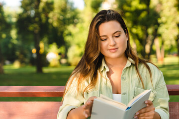 Fototapeta premium Caucasian young woman student freelancer reading book novel, doing homework, preparing for test exam lesson sitting in college university park bench