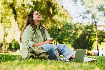 Naklejka na ściany i meble Smiling relaxing caucasian freelancer student e-learning remotely on distance, using laptop, doing homework, project, preparing for exam test in college university, working and drinking coffee in park