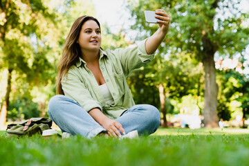Smiling caucasian student freelancer having video call conversation on smart phone, taking selfie, vlogging, blogging on mobile application, social media posting in park