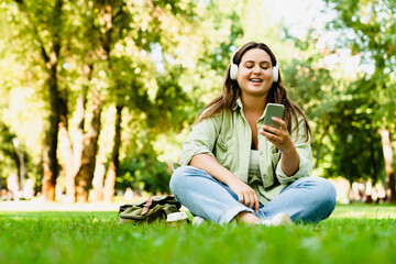 Relaxing caucasian young freelancer teenager student woman listening to the music song singer playlist radio podcast sitting in lotus position in musical mobile application in park