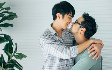 Portrait Gay LGBT sweet Asian couple wearing pajamas, smiling, kissing and hugging with happiness and love, sitting in living room at home with comfortable, cute natural posing in Lifestyle Concept.