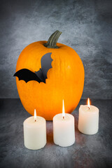 Fresh orange pumpkin with black paper bat and three burning candles on gray surface