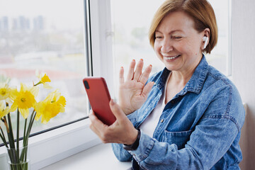 Smiling mature blonde woman making video call with smartphone, gesture hi, having pleasant conversation, wearing blue denim jeans shirt and modern headset using wireless internet connection at home