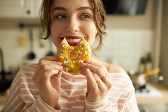 Crop Shot Of Beautiful Young Teenage Girl With Bright Make Up And Red Lipstick On Lips Holding Bitten Doughnut Near Her Mouth With Cute Smile Showing White Healthy Teeth, Looking Aside With Pure Joy