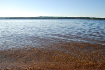 Small waves on a wide river. Summer day, clear water of light brown color from the sand at the bottom of the reservoir. The surface of the water with low waves rushing to the sandy shore.