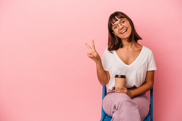 Young mixed race woman sitting on a chair holding a coffee isolated on pink background joyful and carefree showing a peace symbol with fingers.