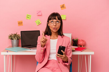 Young mixed race woman preparing a exam for university holding a mobile phone isolated on pink background pointing upside with opened mouth.