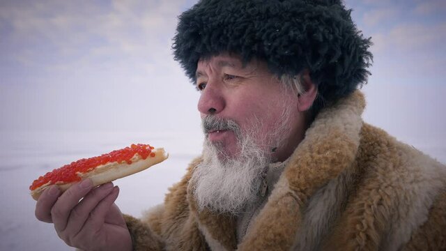 Side view close-up old northern indigenous man eating red caviar sandwich outdoors. Senior Asian guy enjoying delicious taste of healthful food resting at white ice landscape