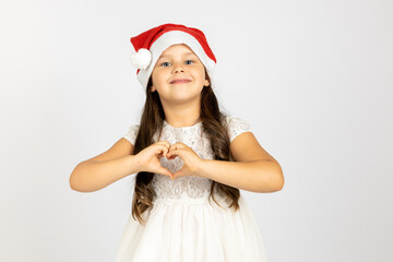 portrait of smiling, beautiful girl in white dress, wearing red Santa Claus hat shows heart sign from fingers isolated on white background.