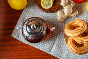 A kettle of strong hot tea on a napkin next to lemon, ginger and jam and donuts.