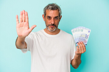 Middle age caucasian man holding bills isolated on blue background  standing with outstretched hand showing stop sign, preventing you.