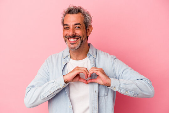 Middle age caucasian man isolated on pink background  smiling and showing a heart shape with hands.
