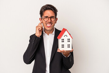 Young mixed race business man holding a toy house isolated on white background covering ears with hands.