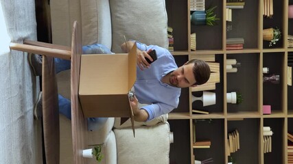 Unpacking parcel. Vertical video young man sit on couch open postal package got by courier service hold phone check web order. Male addressee contact sender by cell text message of receiving delivery - Powered by Adobe
