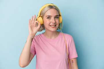 young caucasian woman listening to music isolated on blue background cheerful and confident showing ok gesture.
