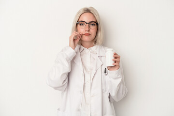 Young pharmacist woman holding pills isolated on white background with fingers on lips keeping a secret.