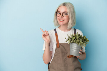 Young gardener caucasian woman holding a plant isolated on blue background smiling and pointing...