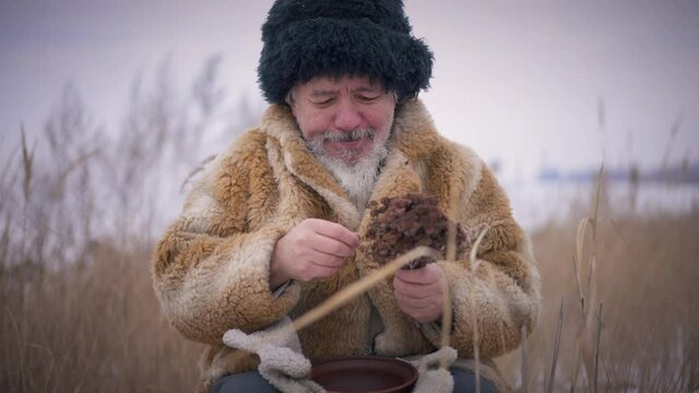 Front view smiling indigenous shaman choosing withered flowers outdoors on cold winter day. Positive confident senior old man selecting plants for healing in slow motion