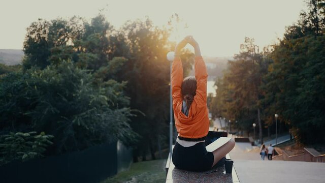 The tired girl stretches after a long period of online work. Dressed in a bright orange blouse, hair caught in a bun, positioned on the park strip. 4k concept