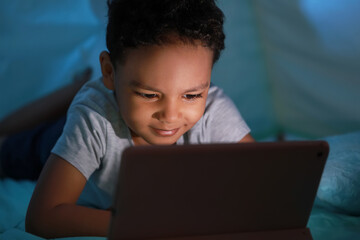 Little African-American boy watching cartoons on tablet computer in play tent at night