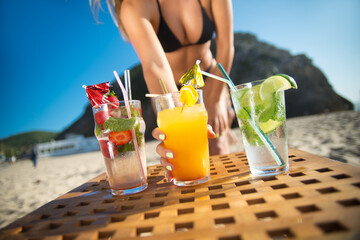 Close-up of woman taking cocktail. Multicolored fancy drinks in glasses on table, woman in black swimsuit holding glass. Party, outdoor activity, beverages concept