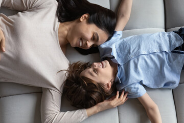 Obraz na płótnie Canvas Happy cute boy and Asian mom relaxing on couch, talking, smiling, laughing, enjoying leisure time at home together. Cheerful mother and little son resting on comfortable couch. Top view