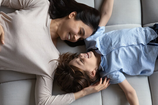 Happy cute boy and Asian mom relaxing on couch, talking, smiling, laughing, enjoying leisure time at home together. Cheerful mother and little son resting on comfortable couch. Top view