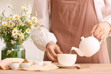 Fototapeta na wymiar Woman pouring fresh chamomile tea from teapot into cup on table