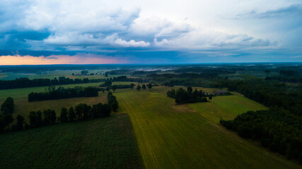 Fototapeta premium Aerial view of the sunset over green fields and forest. Heavy clouds in the sky. 