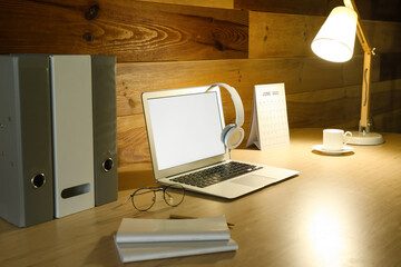 Modern workplace with laptop, headphones and glowing lamp near wooden wall
