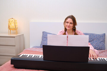 Young woman in pink pajamas playing notes on home bed. Musician playing a melody on a digital piano...