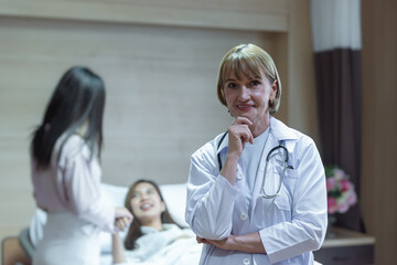Fototapeta premium Caucasian senior woman doctor smiling while holding hand on chin and standing with background have the relatives visiting the patients who were treated improved.