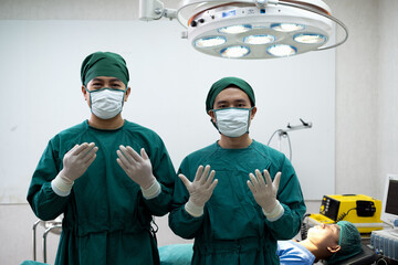 Two Doctors in operating room wearing green mask surgical gown surgeon hair cap latex glove one stand in front of bed and electrical defibrillator other.