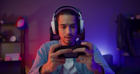 Happy asia man gamer wear headphone competition play video game online with smartphone colorful neon light in living room at night modern house. Esport streaming game online, Home quarantine activity.