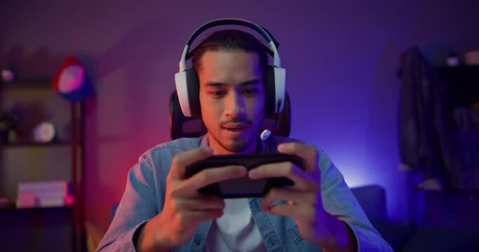 Happy asia man gamer wear headphone competition play video game online with smartphone colorful neon light in living room at night modern house. Esport streaming game online, Home quarantine activity.