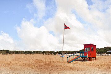 Fototapeta na wymiar Red wooden cabin on the beach for coastguard. Coast guard cabin in Ostende (coast Argentina). Dangerous sea flag on a sunny beach day