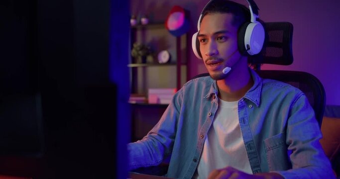Happy asia guy gamer wear headphone participation play video game colorful neon lights computer in living room at night modern house. Esport streaming game online, Home quarantine activity concept.