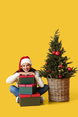 Young woman with gifts and Christmas tree on yellow background