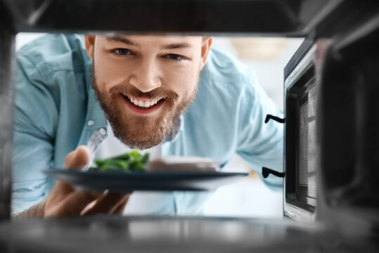 Young man putting plate with food into microwave oven, view from inside