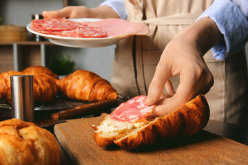 Woman preparing tasty croissant sandwich with meat at kitchen table