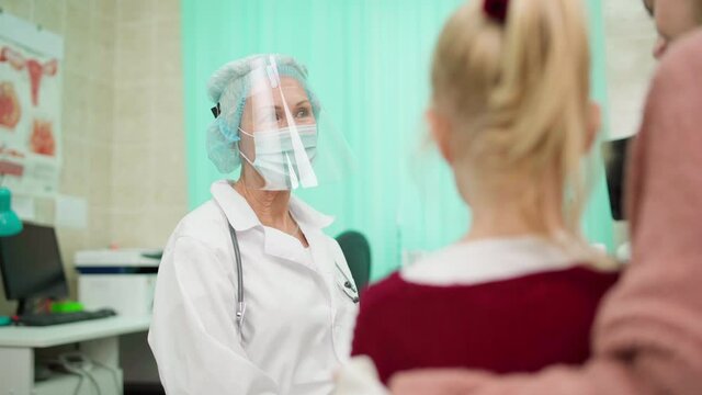 Zoom in rack focus shot of senior female doctor in protective face shield, mask and white coat talking to mother visiting pediatrician with little daughter. Mom hugging kid patient supportively