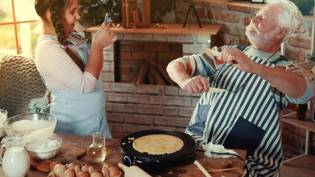 Cute little girl takes photos on the phone of a cheerful grandfather while baking pancakes. Old man poses for a photo while cooking