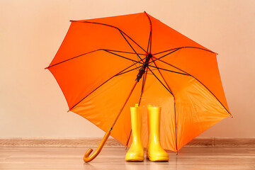 Pair of rubber boots with umbrella on floor against color wall