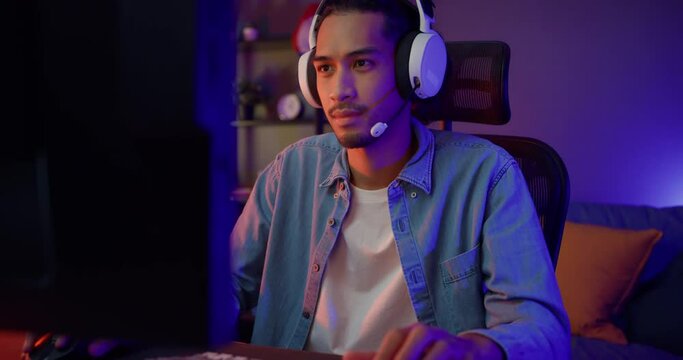 Happy asia guy gamer wear headphone participation play video game colorful neon lights computer in living room at night modern house. Esport streaming game online, Home quarantine activity concept.