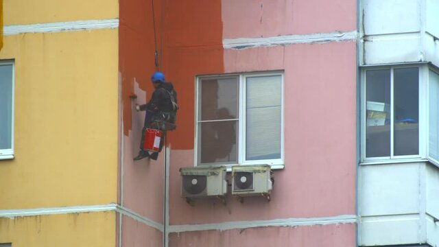 paint pink color high rise building on street on cables in helmets. man on roll wall. facade worker with brush. safety construction with lift rope belt in city update. Reconstruction residential town
