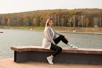 a young beautiful girl in a gray jacket is sitting on the pier of the river autumn city park