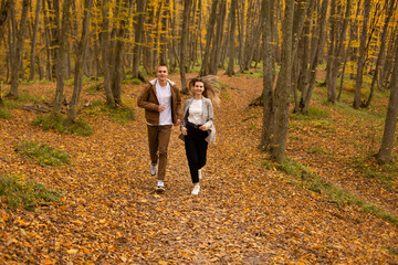 Fototapeta premium a young handsome guy in an autumn beige jacket runs through the autumn city forest park with a young beautiful girl in a gray jacket