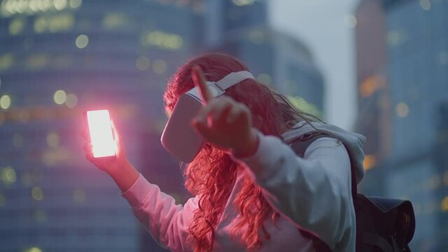 Curly haired young woman in virtual reality glasses looks around and highlights with pink smartphone screen standing against blurry modern financial center building
